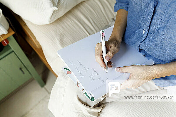 Woman writing in book sitting on bed at home