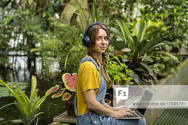 Young woman sitting with laptop listening music through wireless headphones in garden