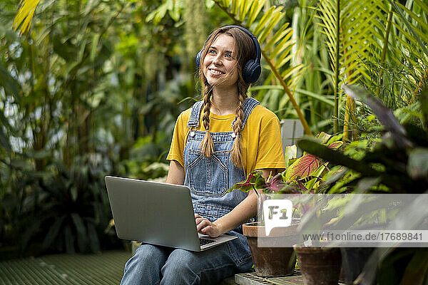 Happy woman with laptop listening music through wireless headphones in garden