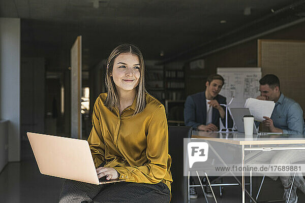 Businesswoman using laptop in office with colleagues in background