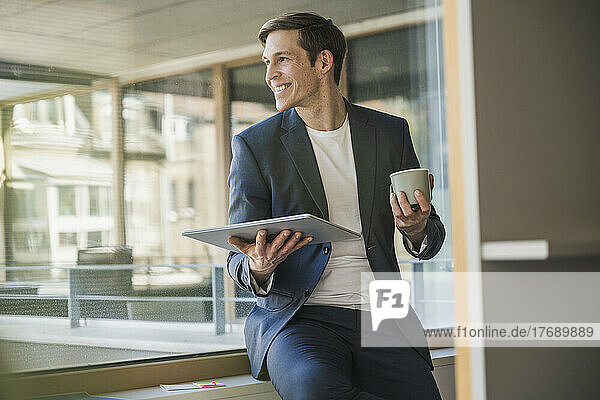 Confident businessman holding tablet and coffee mug at the window in office