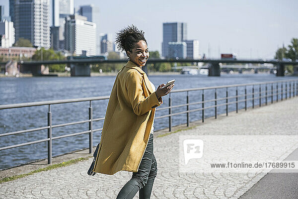 Lächelnde Geschäftsfrau mit Handy spaziert an sonnigem Tag am Main entlang