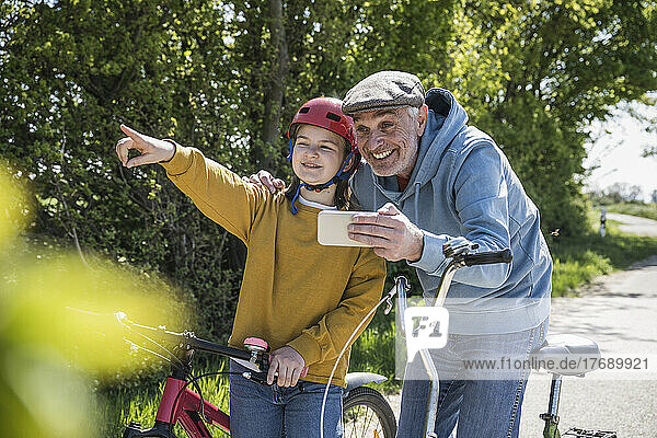 Happy senior man holding mobile phone standing with granddaughter pointing away