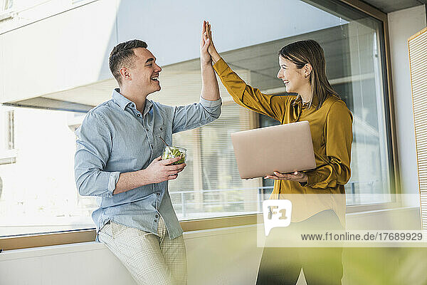 Businessman and businesswoman with laptop high fiving at the window in office