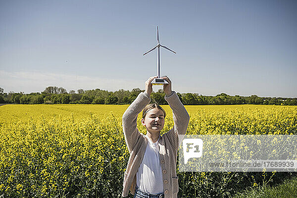 Lächelndes Mädchen mit geschlossenen Augen hält ein Modell einer Windkraftanlage  die auf dem Feld steht