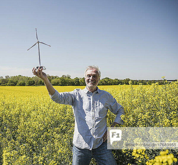 Glücklicher Mann hält Modell einer Windkraftanlage und steht mit der Hand auf der Hüfte auf dem Feld