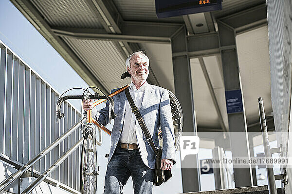 Lächelnder Geschäftsmann mit Fahrrad auf der Schulter  der am Bahnhof spaziert