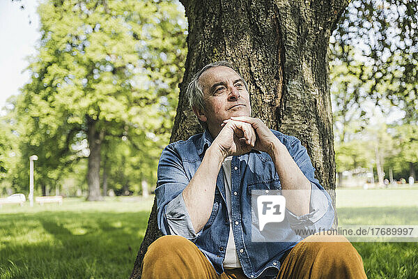 Thoughtful senior man with hand on chin sitting in front of tree at park