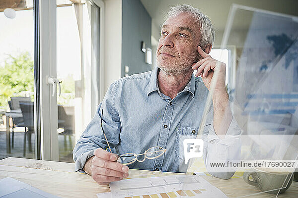 Thoughtful senior freelancer with transparent screen sitting at table