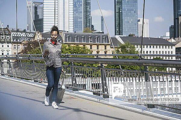 Junge Frau benutzt Smartphone und geht mit Gepäck auf Brücke