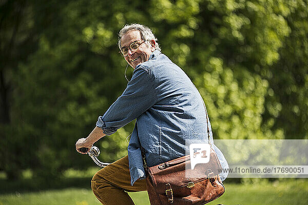 Happy senior man with crossbody bag riding bicycle at park