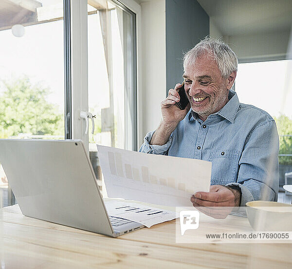 Happy freelancer with documents talking on smart phone at home