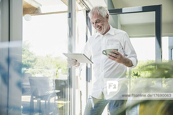 Happy senior man with tablet PC and coffee cup standing by glass window