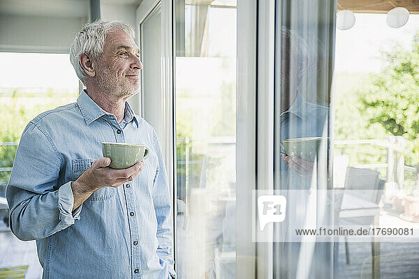 Happy senior man holding coffee cup looking out through window