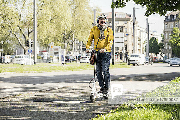 Smiling senior man wearing wireless headphones riding on electric push scooter