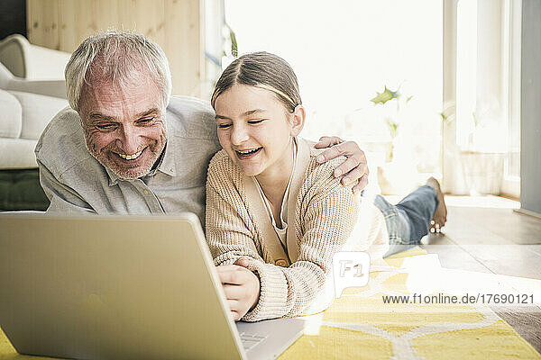 Glücklicher älterer Mann mit Enkelin  der zu Hause auf dem Teppich im Wohnzimmer liegt und einen Laptop benutzt