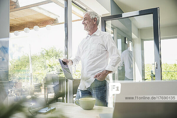 Lächelnder älterer Mann mit Tablet-PC vor Glasfenster zu Hause