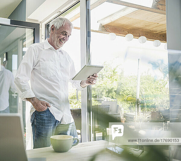Happy senior man using tablet PC standing by glass window at home