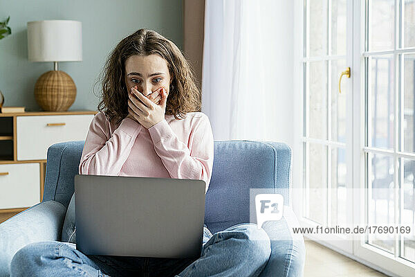Young freelancer covering mouth with hands looking at laptop