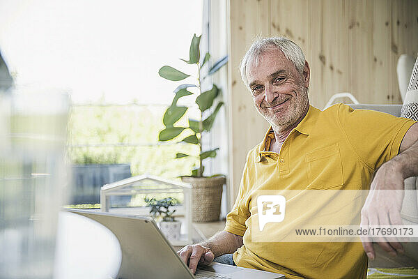 Smiling senior man with laptop sitting in living room at home