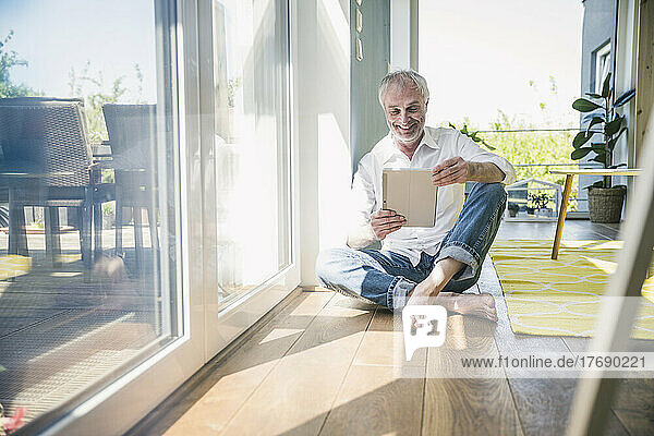 Happy senior man using tablet PC sitting on floor in living room at home