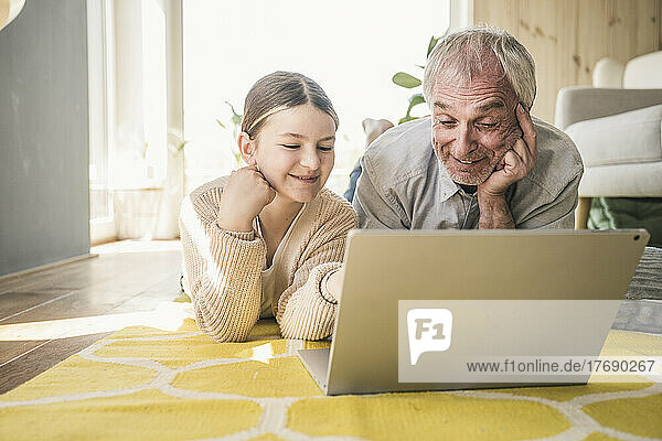 Lächelnder älterer Mann mit Enkelin  der zu Hause auf dem Teppich liegt und einen Laptop benutzt
