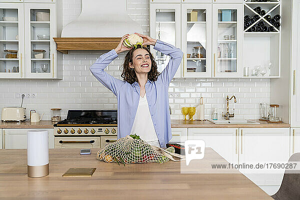 Smiling woman holding cauliflower on head in kitchen