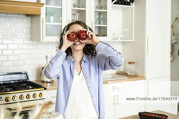 Smiling woman covering eyes with apples in kitchen