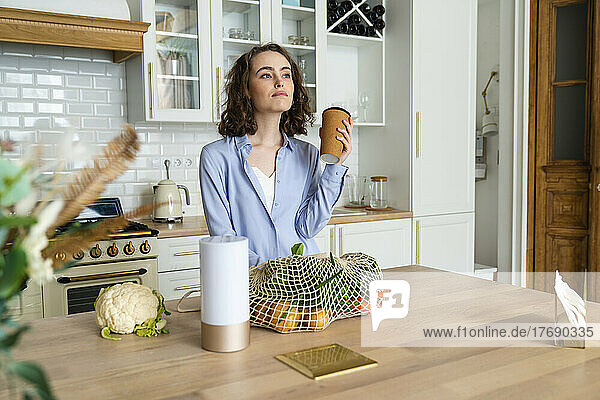 Thoughtful woman with disposable coffee cup in kitchen