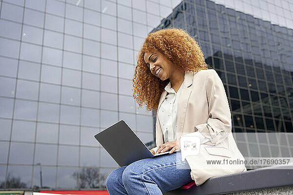 Smiling businesswoman working on laptop outside office building