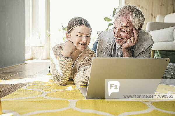 Lächelnder älterer Mann mit Enkelin  der zu Hause auf dem Teppich im Wohnzimmer liegt und einen Laptop benutzt