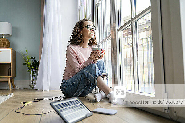 Junge Geschäftsfrau mit Kaffeetasse schaut zu Hause durch das Fenster