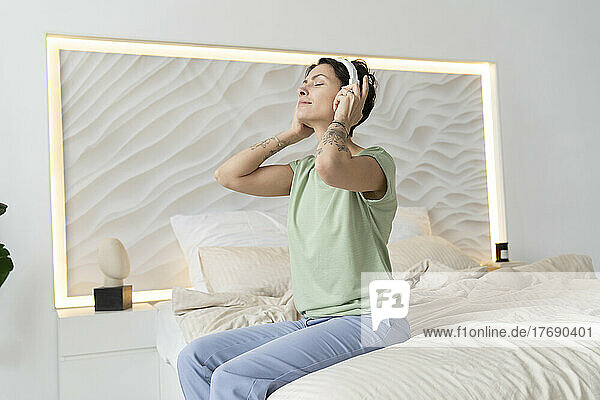 Woman with eyes closed enjoying music listening through wireless headphones sitting on bed at home