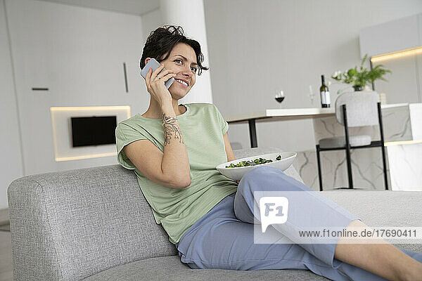 Smiling woman holding salad bowl talking on mobile phone at home