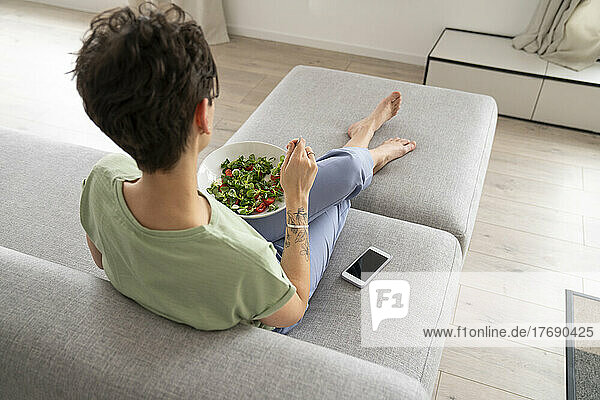 Frau isst Salat und sitzt zu Hause mit dem Handy auf dem Sofa