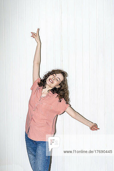 Happy young woman with arms raised dancing in front of white wall