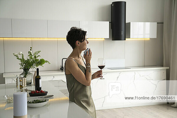 Happy woman holding wineglass talking through mobile phone speaker in kitchen