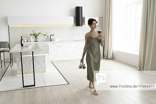 Woman holding high heels and drinking wine walking in kitchen at home