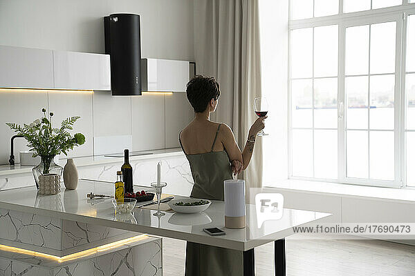 Woman holding wineglass looking through window in kitchen at home