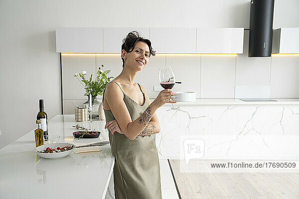 Smiling woman holding wineglass leaning on kitchen island at home