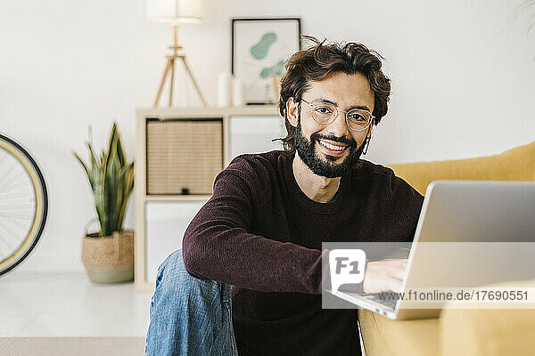 Smiling man with laptop sitting by sofa at home