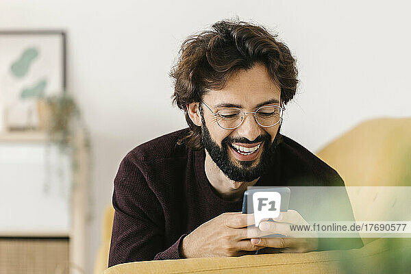 Lächelnder bärtiger Mann  der zu Hause auf dem Sofa sein Mobiltelefon benutzt
