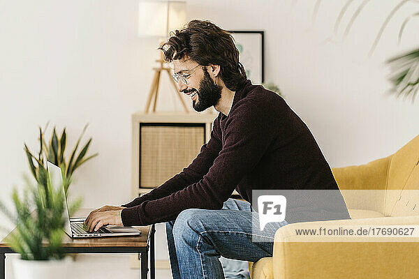 Smiling bearded man using laptop sitting on sofa at home