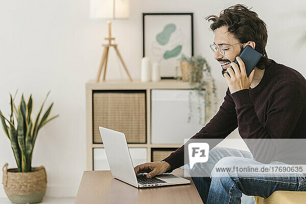 Happy man with laptop talking on mobile phone at home