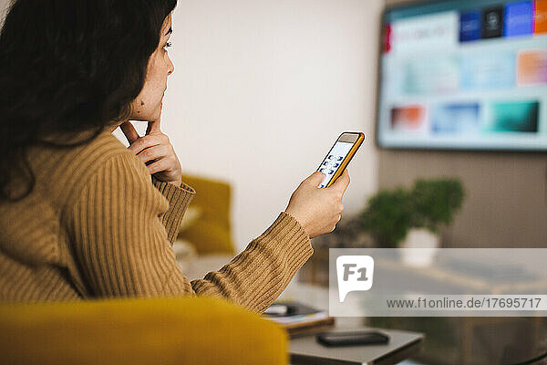 Seitenansicht einer jungen Frau  die zu Hause ihren Fernseher über eine mobile App steuert