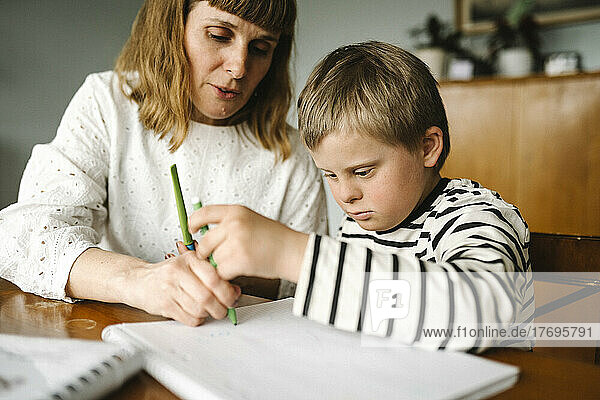 Junge mit Behinderung  der von seiner Mutter unterstützt wird  während er zu Hause in ein Buch zeichnet