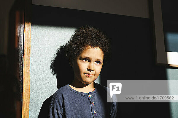 Boy looking away standing in sunlight against wall at home