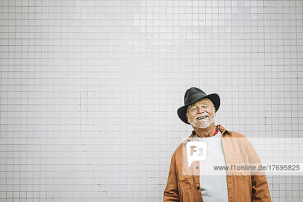 Porträt eines glücklichen älteren Mannes mit Hut  der an einer weißen Wand steht