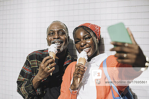 Smiling man taking selfie with father eating ice cream during sunny day
