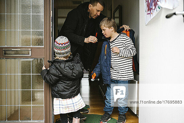 Father helping son with disability to wear jacket by daughter standing at doorway